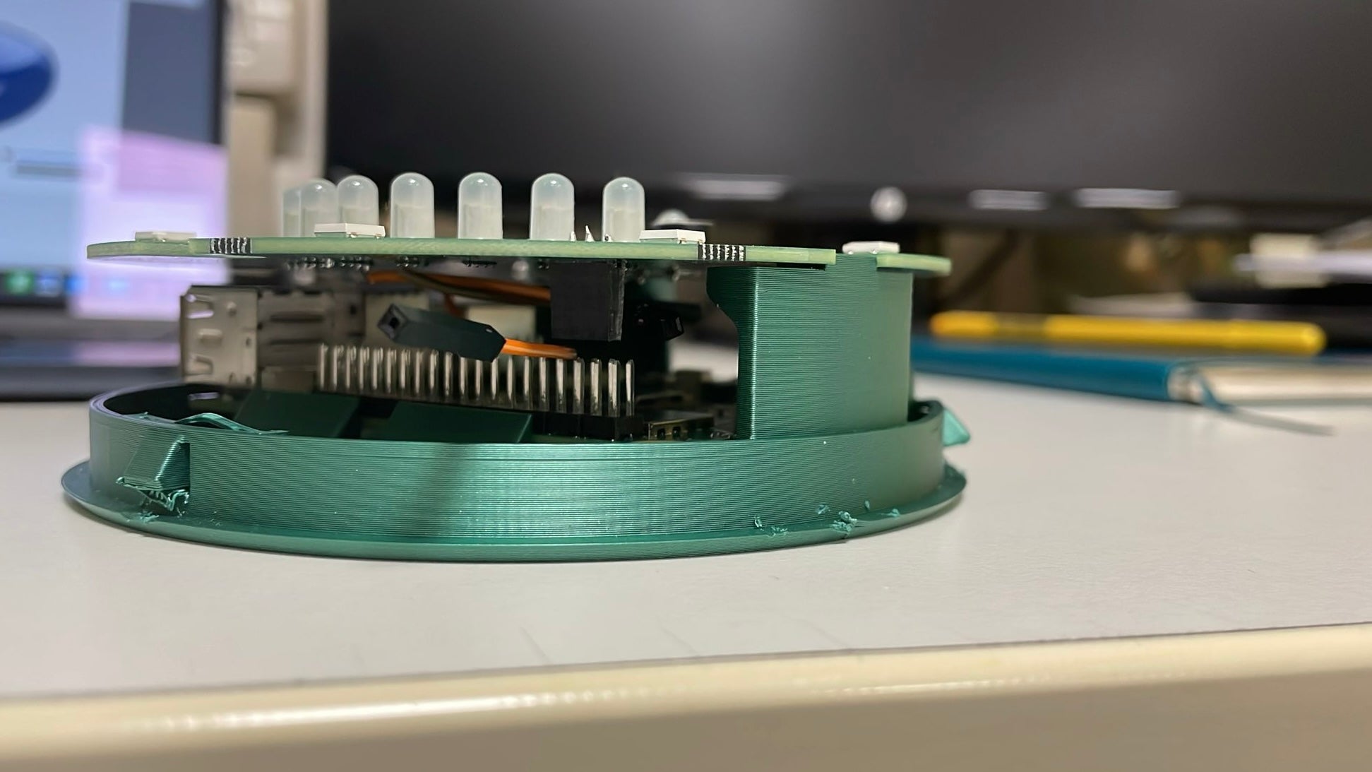 Primo piano di un prototipo composto da una base verde stampata in 3D e componenti elettroniche, posizionato su una scrivania bianca, monitor e agenda sullo sfondo