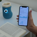 Smartphone che mostra l'app Drinky con grafici di idratazione e parametri vitali, accanto al bicchiere intelligente azzurro con sensore LED circolare illuminato su un tavolo