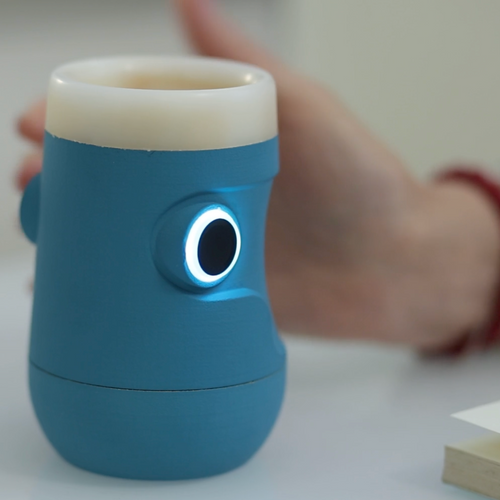 Bicchiere intelligente Drinky azzurro e bianco in primo piano su un tavolo bianco, con sensore LED circolare bianco acceso, mano e libro aperto sullo sfondo.