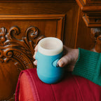 Bicchiere intelligente Drinky azzurro e bianco, vista laterale in ambiente domestico, impugnato da una mano e posato sul bracciolo di un divano.
