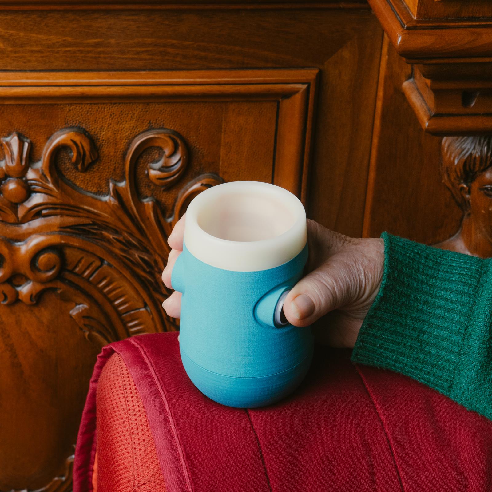 Bicchiere intelligente Drinky azzurro e bianco, vista laterale in ambiente domestico, impugnato da una mano e posato sul bracciolo di un divano.