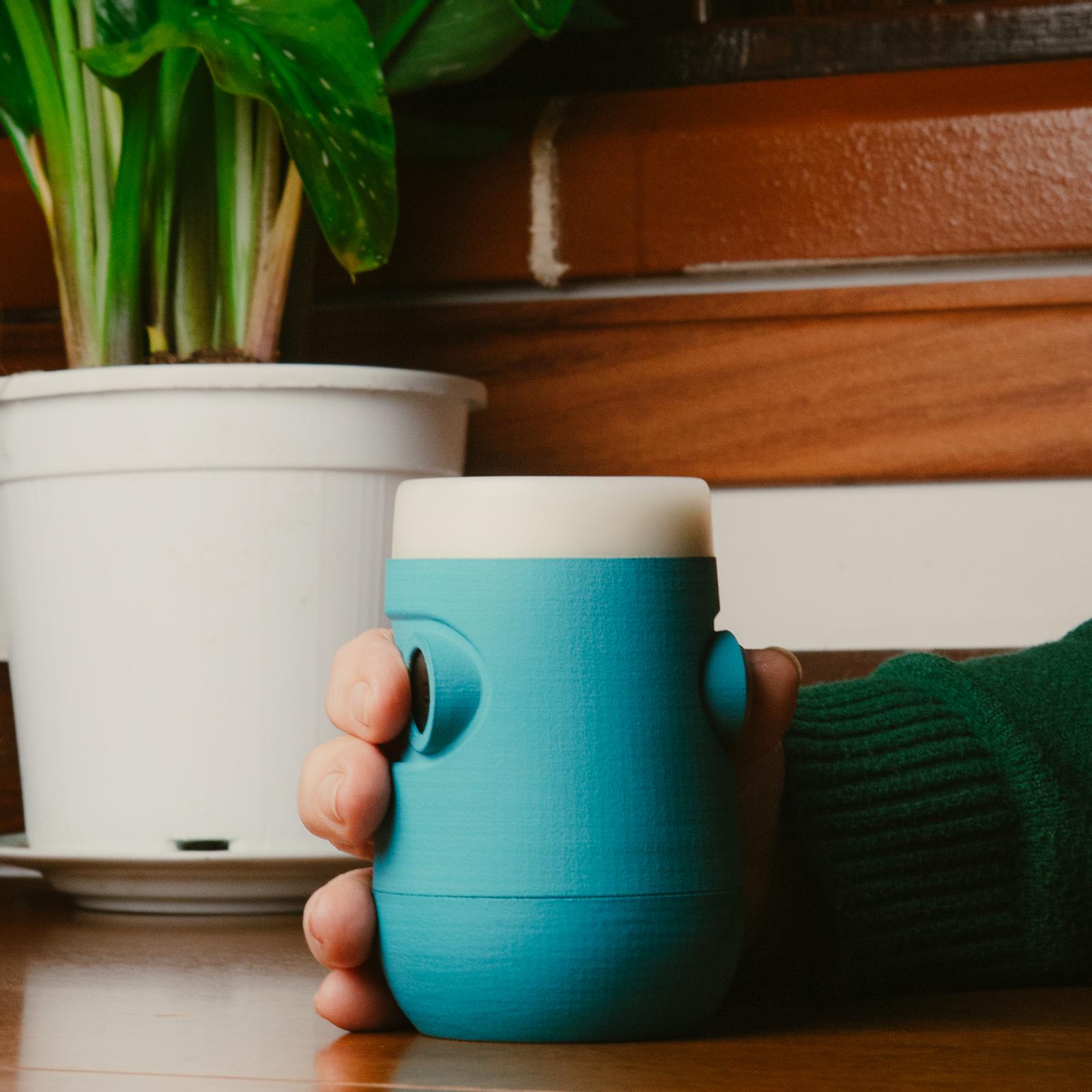 Bicchiere intelligente Drinky azzurro e bianco, vista frontale in ambiente domestico, impugnato da una mano e posato su un tavolo.